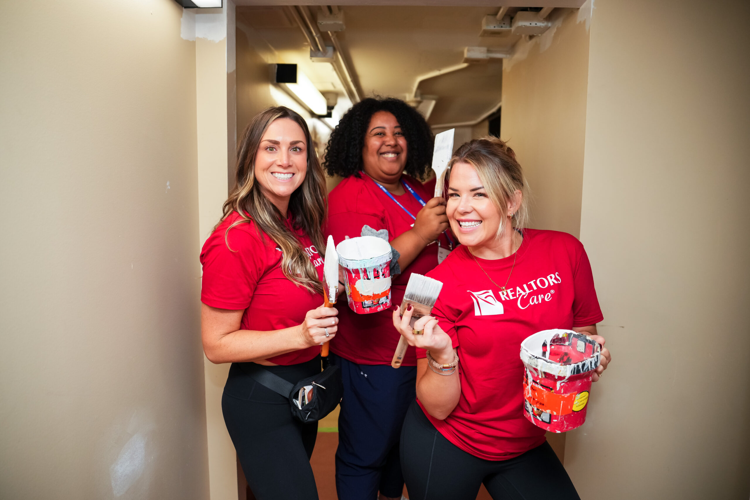 Three REALTORS Care volunteers holding paintbrushes and paint cans in a hallway.